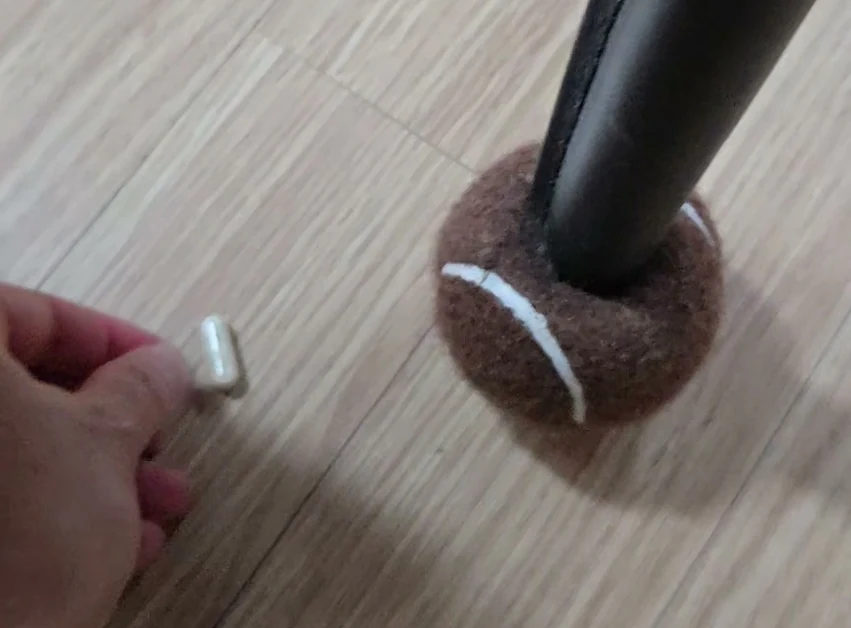 마룻바닥에 놓인 흰 캡슐 한 알과 그쪽으로 뻗은 손끝, 옆에 의자 다리 덮개